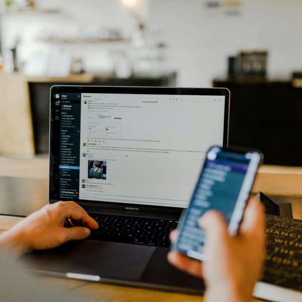 Eine nicht sichtbare Person sitzt an einem Tisch und hat in der einen Hand ein Smartphone, während die andere Hand einen Laptop bedient. Im Hintergrund ist verschwommen ein helles Büro oder Cafe zu erkennen.