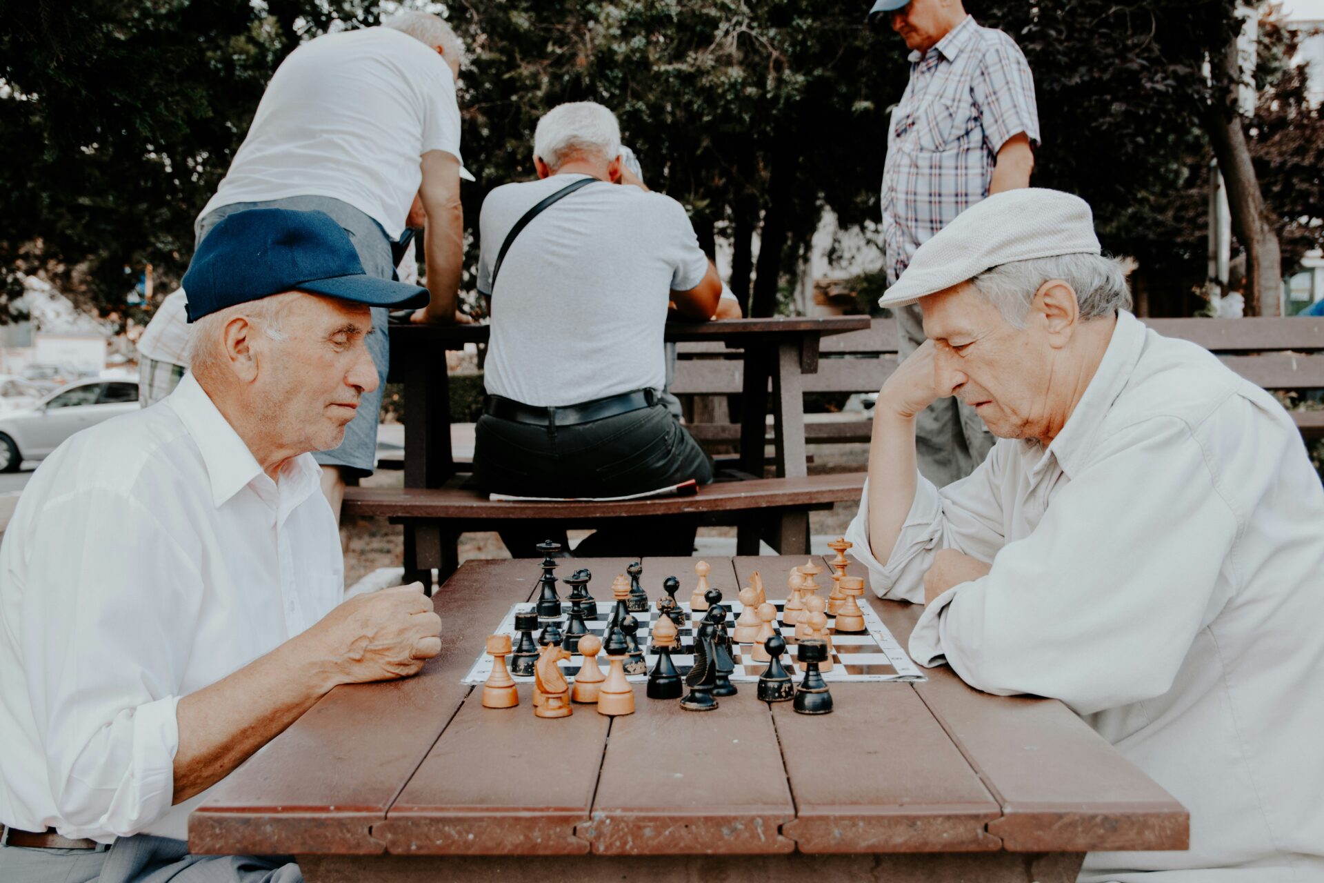Zwei ältere Männer sitzen an einem Tisch im Park und sind in eine Partie Schach vertieft. Im Hintergrund sind weitere Männer zu erkennen, die sich um einen zweiten Tisch versammeln.
