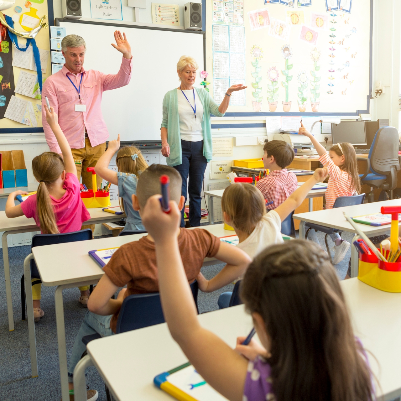 Ein Bild aus einem Klassenzimmer. Die Kinder sind von hinten zu sehen, vorne steht eine Frau und ein Mann. Manche heben die Hände.